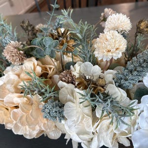 Puede incluir: Primer plano de un arreglo floral con una variedad de flores artificiales en tonos crema, beige y verde apagado. El arreglo incluye hortensias, peque&ntilde;as flores redondas y ramitas de vegetaci&oacute;n, creando un aspecto natural y texturizado.
