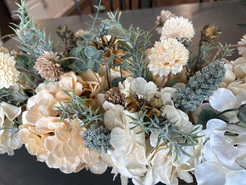 Puede incluir: Primer plano de un arreglo floral con una variedad de flores artificiales en tonos crema, beige y verde apagado. El arreglo incluye hortensias, peque&ntilde;as flores redondas y ramitas de vegetaci&oacute;n, creando un aspecto natural y texturizado.