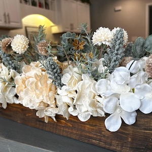 Puede incluir: Centro de mesa rectangular de madera lleno de flores artificiales. El arreglo incluye hortensias blancas, crema y bronceadas, junto con otras plantas y acentos. La caja de madera tiene un acabado marr&oacute;n oscuro.