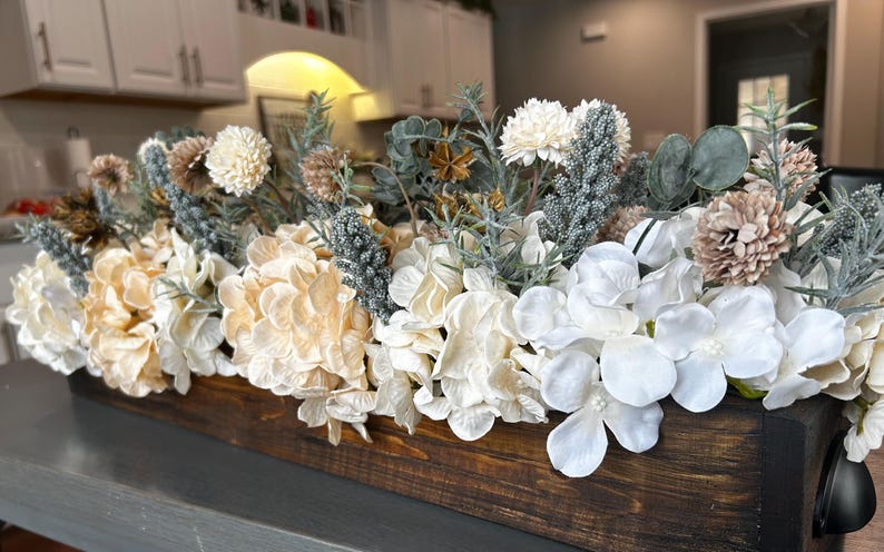 Puede incluir: Centro de mesa rectangular de madera lleno de flores artificiales. El arreglo incluye hortensias blancas, crema y bronceadas, junto con otras plantas y acentos. La caja de madera tiene un acabado marr&oacute;n oscuro.