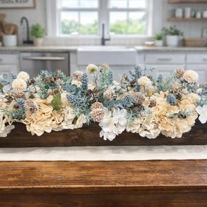 Puede incluir: Centro de mesa rectangular de madera, lleno de flores y vegetaci&oacute;n artificiales crema, blancas y azules. El arreglo incluye hortensias, pi&ntilde;as, sobre un camino de mesa blanco.