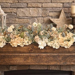 Puede incluir: Un centro de mesa de madera lleno de flores y vegetaci&oacute;n de color crema y azul claro. El arreglo se encuentra sobre una repisa de madera r&uacute;stica frente a una chimenea de piedra. Velas decorativas, una estrella y una linterna completan la exhibici&oacute;n.