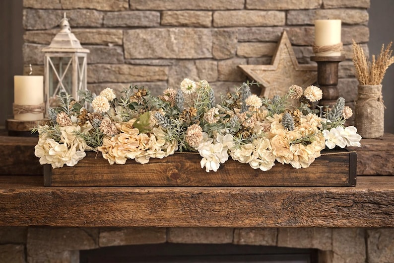 Puede incluir: Un centro de mesa de madera lleno de flores y vegetaci&oacute;n de color crema y azul claro. El arreglo se encuentra sobre una repisa de madera r&uacute;stica frente a una chimenea de piedra. Velas decorativas, una estrella y una linterna completan la exhibici&oacute;n.