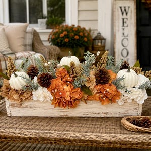 May include: A rectangular, distressed white wooden planter box filled with a seasonal arrangement of white pumpkins, brown pinecones, and orange and white hydrangea flowers, accented with green foliage and tall, textured golden-brown stems.