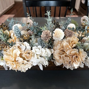 Puede incluir: Un centro de mesa floral con una variedad de flores artificiales en tonos crema, beige y verde apagado. El arreglo incluye hortensias, pompones y otros elementos decorativos, creando una est&eacute;tica natural y elegante. Perfecto para la decoraci&oacute;n del hogar.