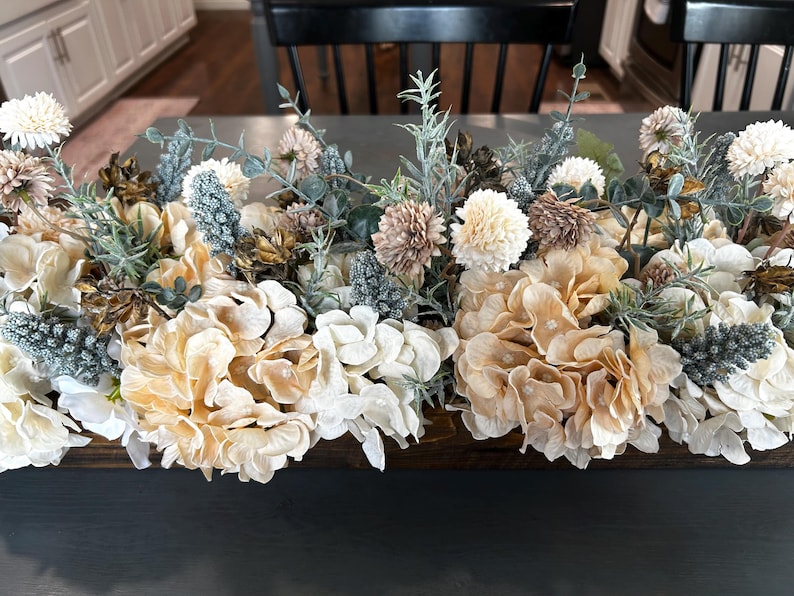 Puede incluir: Un centro de mesa floral con una variedad de flores artificiales en tonos crema, beige y verde apagado. El arreglo incluye hortensias, pompones y otros elementos decorativos, creando una est&eacute;tica natural y elegante. Perfecto para la decoraci&oacute;n del hogar.
