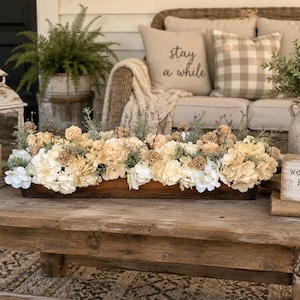 May include: A long, rectangular wooden trough centerpiece filled with cream and white artificial hydrangea flowers, small tan pom-pom blooms, and green sprigs. A small wooden sign with the text "welcome home" and a heart icon sits nearby.