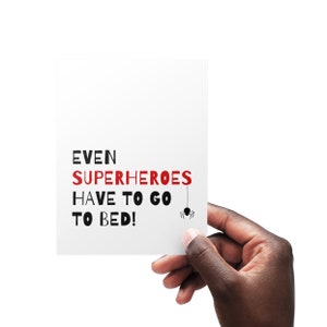 May include: White card with black text that reads "EVEN SUPERHEROES HAVE TO GO TO BED!" with a black spider hanging from the word "GO".
