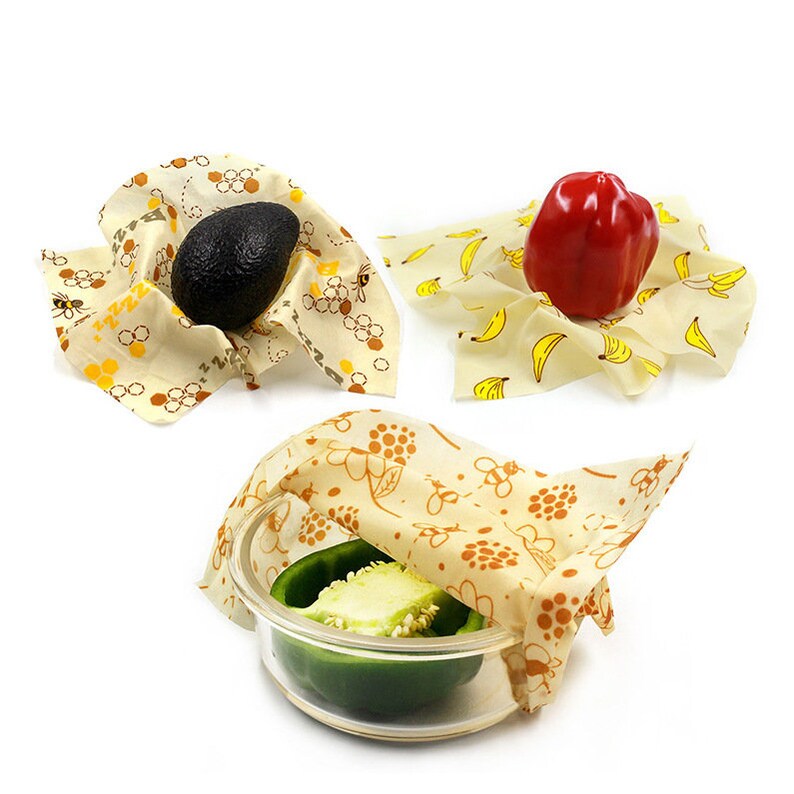 K&ouml;nnte beinhalten: Drei wiederverwendbare Bienenwachst&uuml;cher mit verschiedenen Mustern. Ein Tuch bedeckt einen gr&uuml;nen Paprika in einer Glassch&uuml;ssel, ein anderes bedeckt eine ganze Avocado und das dritte bedeckt einen roten Paprika.