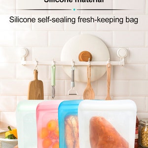 K&ouml;nnte beinhalten: Drei selbstdichtende Frischhaltebeutel aus Silikon in Wei&szlig;, Rosa und Blau. Die Beutel sind mit verschiedenen Lebensmitteln gef&uuml;llt, darunter Obst, Brot und Fleisch. Die Beutel sind mit Messungen in Millilitern gekennzeichnet.