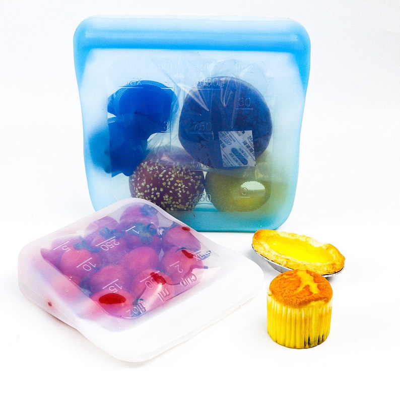 K&ouml;nnte beinhalten: Zwei Silikon-Lebensmittelbeh&auml;lter, einer blau und einer transparent, mit verschiedenen Snacks gef&uuml;llt. Der blaue Beh&auml;lter enth&auml;lt einen Donut, ein Geb&auml;ck und eine Frucht. Der transparente Beh&auml;lter enth&auml;lt kleine rote Tomaten. Die Beh&auml;lter befinden sich auf einer wei&szlig;en Oberfl&auml;che.
