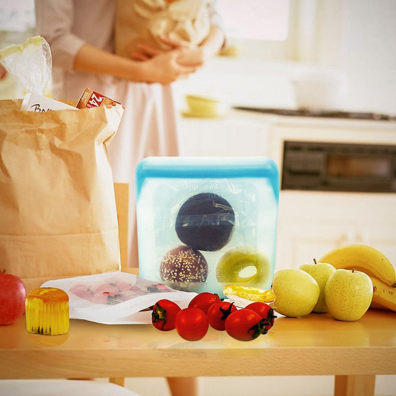 K&ouml;nnte beinhalten: Eine blaue Silikon-Aufbewahrungstasche f&uuml;r Lebensmittel mit verschiedenen Snacks, darunter ein Donut, ein Geb&auml;ck und eine Frucht. Die Tasche liegt auf einem Holztisch mit verschiedenen Fr&uuml;chten und Gem&uuml;se drumherum.
