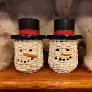 May include: Two white wicker snowman ornaments with black top hats and red ribbon accents. The snowmen have black eyes and a carrot nose.