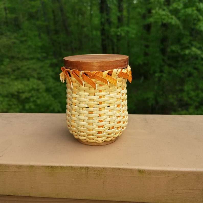May include: A woven basket with a wooden lid and a decorative orange trim. The basket is made of natural materials and has a light brown color.