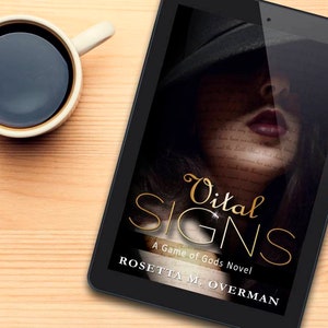 May include: A digital tablet displays the book cover for "Vital Signs: A Game of Gods Novel" by Rosetta M. Overman. The cover features a woman's face with the title in gold script. A cup of coffee sits to the left on a light wood surface.