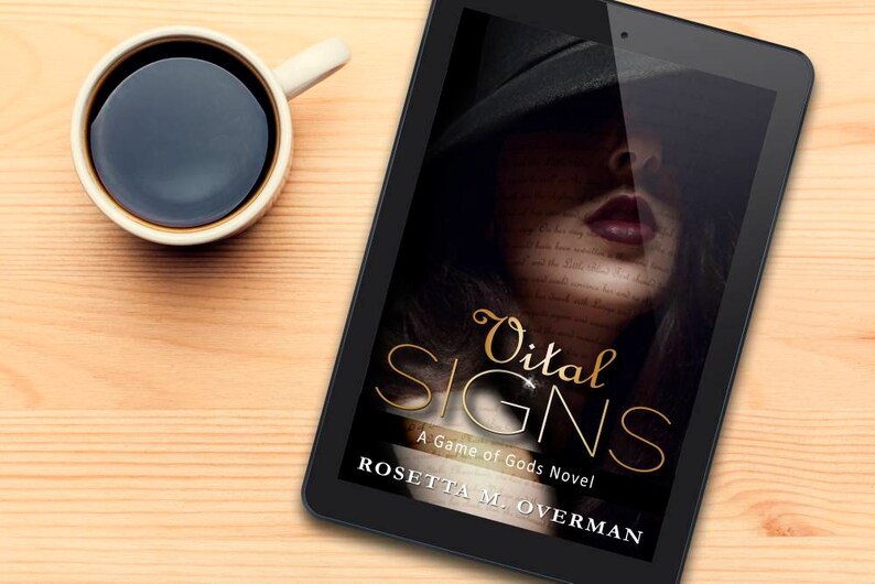 May include: A digital tablet displays the book cover for "Vital Signs: A Game of Gods Novel" by Rosetta M. Overman. The cover features a woman's face with the title in gold script. A cup of coffee sits to the left on a light wood surface.
