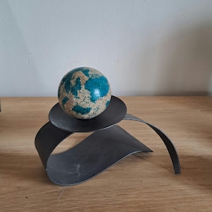 Könnte beinhalten: Ein kugelförmiges Objekt mit einer strukturierten Oberfläche, die gesprenkelte blaue und beige Muster aufweist, ruht auf einem dunkelgrauen Metallständer. Der Ständer besteht aus einem einzigen gebogenen Metallstück, das eine runde Plattform bildet.