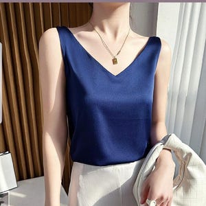 May include: A navy blue sleeveless satin top with a v-neckline. The top is worn with a white skirt and a gold chain necklace with a small rectangular pendant.