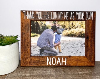 May include: A rectangular wooden picture frame with a dark brown wood grain finish. The top border features the white text THANK YOU FOR LOVING ME AS YOUR OWN and the bottom border features the white text NOAH. The frame holds a photograph.