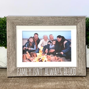 Może przedstawiać: Szara drewniana ramka ze złotym paskiem i zdjęciem rodziny wokół ogniska na plaży. Na dole ramki widnieje napis "FAMILY ISN'T ALWAYS BLOOD".