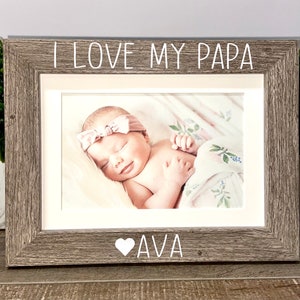 May include: A gray wooden picture frame with a white inscription that reads "I love my Papa" and a heart with the name "Ava" below the photo. The photo shows a baby girl sleeping in a pink and white blanket.
