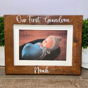 May include: A rectangular wooden picture frame with a dark brown wood grain finish. The top of the frame features the white text "Our First Grandson" and the bottom features the white text "Noah". A photo of a baby in a blue wrap is inside.