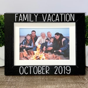 May include: Black picture frame with a white mat and a photo of a family around a campfire on the beach. The frame has the text "FAMILY VACATION OCTOBER 2019" printed on it.