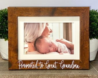 May include: A rectangular wooden picture frame with a dark brown finish and a white mat. The frame features the white text Promoted to Great Grandma printed on the bottom border. Inside the frame is a photograph of an adult holding a baby.