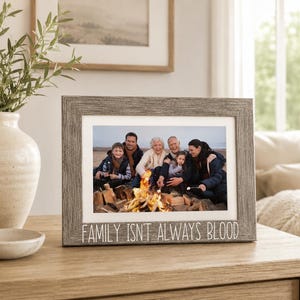 May include: A rectangular gray wood-grain picture frame with a white mat, displaying a photograph of a family gathered around a campfire. The bottom of the frame features the white printed text, "FAMILY ISN'T ALWAYS BLOOD" in a clean, sans-serif font.