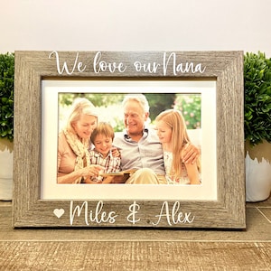 May include: A wood-toned picture frame with a white inscription that reads "We love our Nana" and "Miles & Alex" with a heart symbol. The frame holds a photo of a family with two children, a woman, and a man.