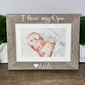 May include: A gray wooden picture frame with a white mat. The frame has the text "I love my Opa" at the top and a heart with the name "AVA" at the bottom. The frame contains a photo of a baby girl sleeping.