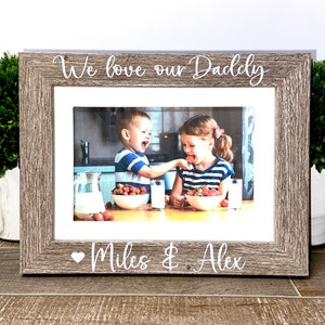 May include: A rustic wood picture frame with a white inscription that reads "We love our Daddy" and a heart with the names "Miles & Alex" below. The frame holds a photo of two children smiling and eating strawberries.
