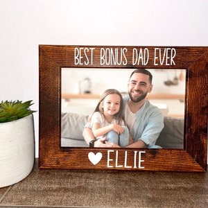 Può includere: Cornice rettangolare in legno marrone scuro con una foto di un uomo e una bambina. La cornice presenta la scritta bianca "BEST BONUS DAD EVER" in alto e "ELLIE" con un cuore in basso.