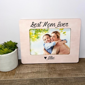 Puede incluir: Marco de fotos de madera rosa con el texto "Best Mom Ever" y un corazón con el nombre "Ellie" debajo de la foto. La foto muestra a una madre y una hija abrazándose.