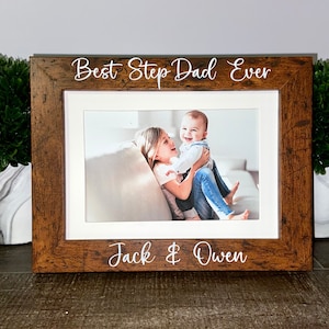 Może przedstawiać: Brązowa drewniana ramka ze złotym paskiem i zdjęciem dwóch dzieci w środku. Ramka ma napis "Best Step Dad Ever" u góry i "Jack & Owen" na dole.