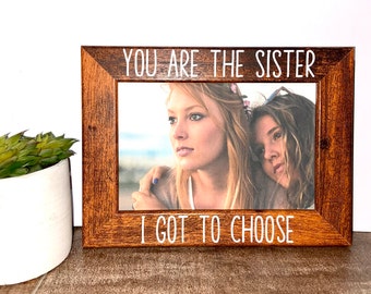 May include: A rectangular wooden picture frame with a dark brown wood grain finish. The top border features the white text YOU ARE THE SISTER and the bottom border features the white text I GOT TO CHOOSE. A photograph of two individuals is inside.
