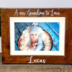 May include: A rectangular dark brown wooden picture frame with a white mat. The top of the frame features the white text A new Grandson to Love, and the bottom features the name Lucas. Inside is a photograph of a baby in a blue hat and wrap.