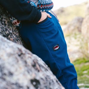 Puede incluir: Primer plano de un pantalón de chándal azul marino con un pequeño parche con el logotipo en la pierna izquierda. El pantalón tiene un corte holgado y bolsillos. Se ve la parte superior de un suéter estampado.