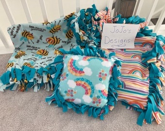 May include: Three turquoise blue fleece tie blankets with different patterns. One blanket has a bee pattern with the text "Bee-lieve in yourself". Another blanket has a rainbow pattern. The third blanket has a rainbow pattern with clouds. A white card with the text "JoJo's Designs" is on top of the rainbow blanket.