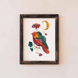 May include: A framed watercolor print featuring a vibrant bird with orange and red plumage, embraced by a green figure. The artwork includes a crescent moon, a flower, and floral accents. The frame is dark brown.