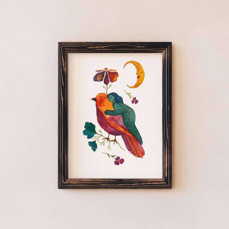May include: A framed watercolor print featuring a vibrant bird with orange and red plumage, embraced by a green figure. The artwork includes a crescent moon, a flower, and floral accents. The frame is dark brown.