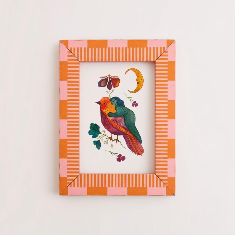 May include: A framed watercolor print featuring a colorful bird with a person hugging it, surrounded by flowers and a crescent moon. The frame has a pink and orange checkered pattern on the edges and a striped design on the sides.