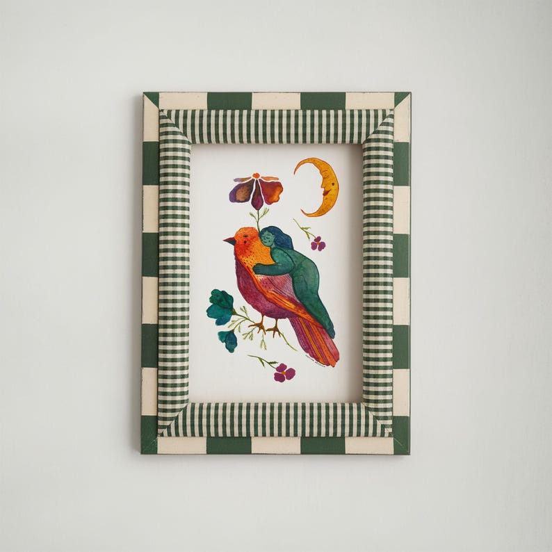 May include: A framed watercolor print featuring a colorful bird with a person hugging it. The artwork includes a flower, moon, and floral accents. The frame has a green and white checkered pattern.