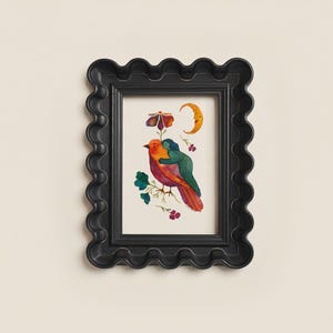 May include: A framed art print featuring a colorful illustration of a bird embraced by a figure, with a flower, crescent moon, and floral accents. The black frame has a wavy, scalloped edge, adding a decorative touch to the artwork.