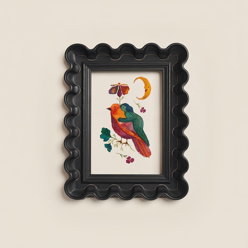 May include: A framed art print featuring a colorful illustration of a bird embraced by a figure, with a flower, crescent moon, and floral accents. The black frame has a wavy, scalloped edge, adding a decorative touch to the artwork.