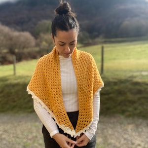 Puede incluir: Un chal de crochet amarillo con un borde de crochet blanco. El chal lo lleva una persona con el pelo oscuro recogido en un moño. La persona está de pie frente a un campo verde con una montaña al fondo.