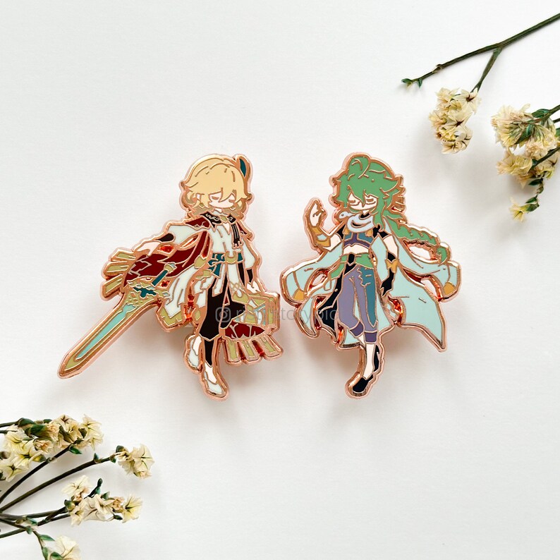 Puede incluir: Dos pines de esmalte de color dorado con personajes de estilo anime. El personaje de la izquierda tiene el pelo rubio y lleva un atuendo rojo y blanco con una espada. El personaje de la derecha tiene el pelo verde y lleva un atuendo azul y verde.