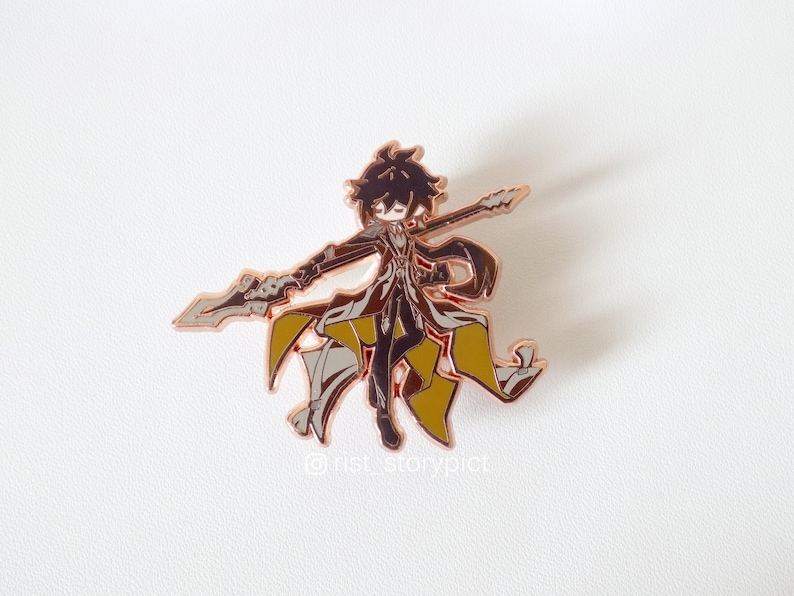 May include: A rose gold enamel pin featuring a cartoon character holding a spear. The character has black hair, a black coat, and yellow accents. The pin is on a white background.