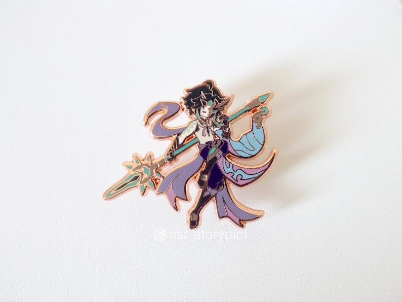 May include: A rose gold enamel pin featuring a character with purple and blue clothing holding a spear with a green and gold star at the end. The character has black hair and blue eyes. The pin has the text "@rist_storypict" at the bottom.