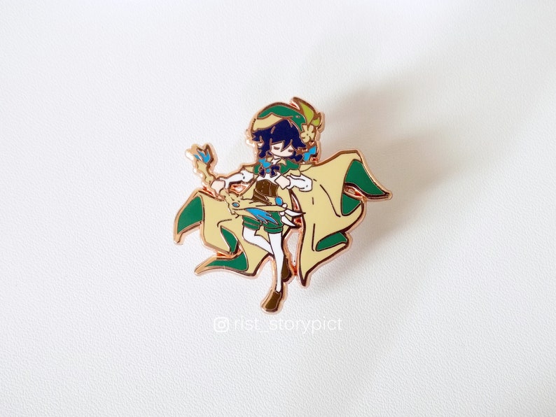 May include: A gold-colored enamel pin featuring a cartoon character with green and yellow clothing, a blue hat, and a green staff. The character has dark blue hair and is holding a staff with a blue and yellow gem. The pin is on a white background.